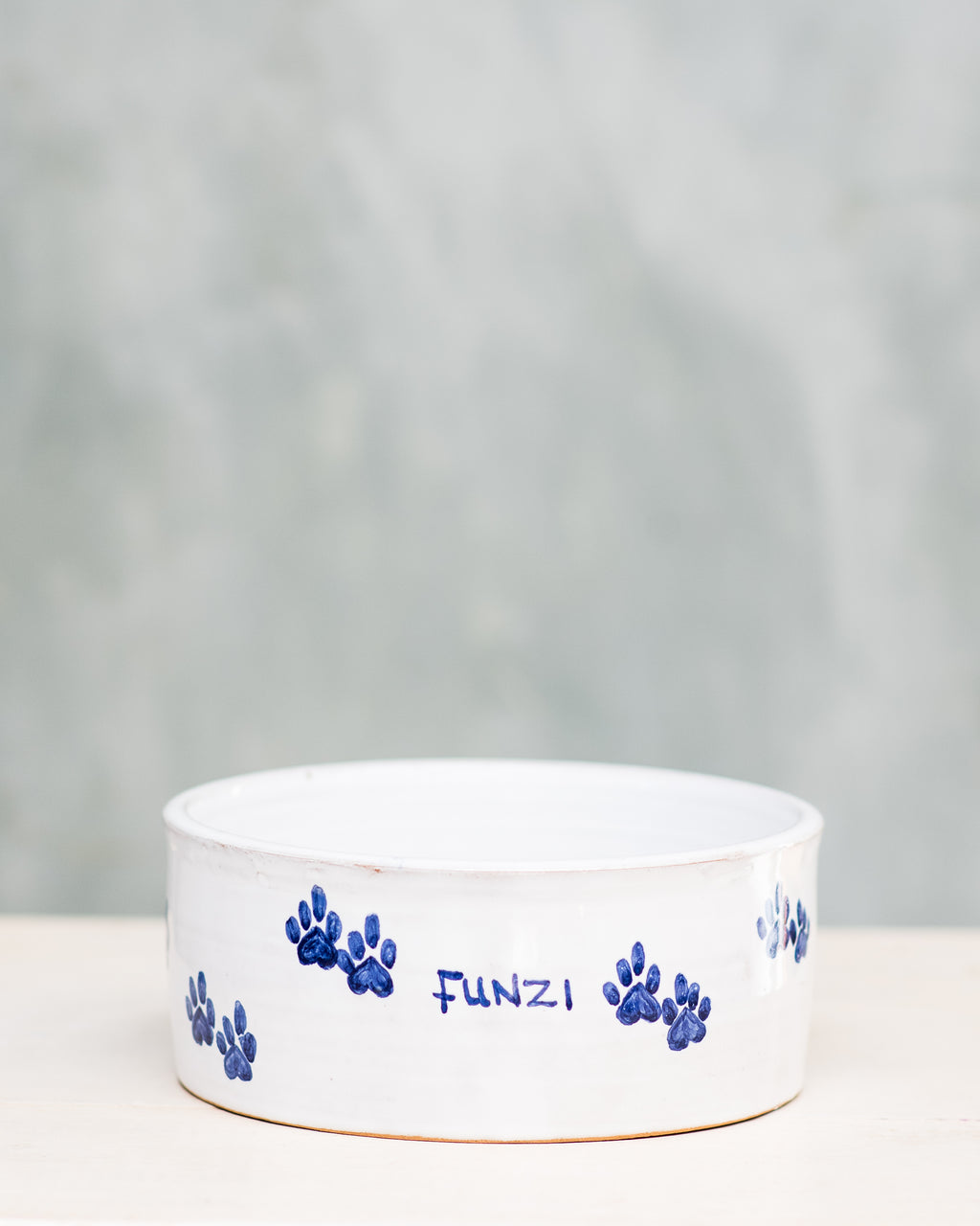 A ceramic dog bowl with blue paw prints around the edge and the name 'Funzi' written on it, placed on a wooden surface with a plain background.