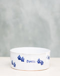 A ceramic dog bowl with blue paw prints around the edge and the name 'Funzi' written on it, placed on a wooden surface with a plain background.