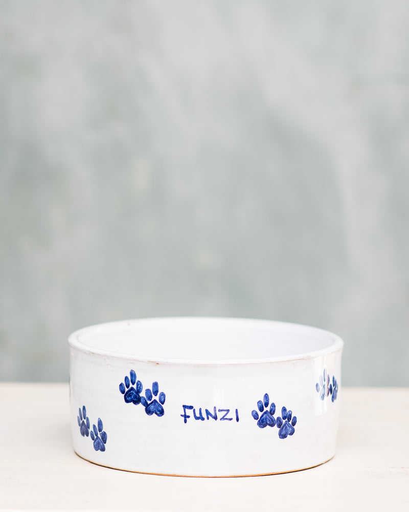 A ceramic dog bowl with blue paw prints around the edge and the name 'Funzi' written on it, placed on a wooden surface with a plain background.