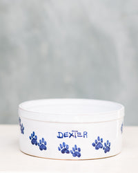A ceramic dog bowl with blue paw prints around the edge and the name 'DEXTER' written on it, placed on a wooden surface with a plain background.