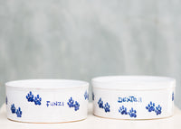 two ceramic dog bowls with blue paw prints around the edge and the name 'Funzi'  'Dexter' written on it, placed on a wooden surface with a plain background.